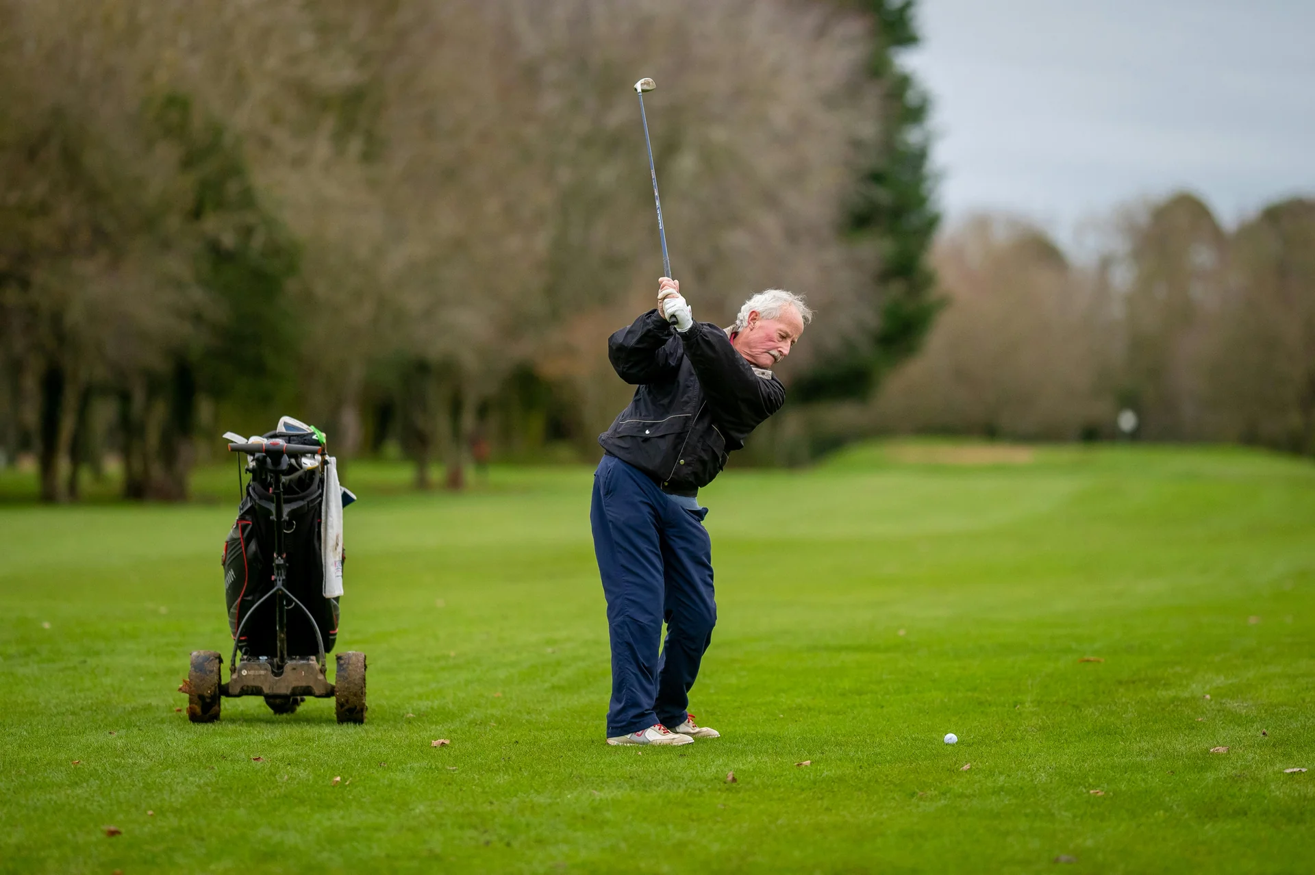 A senior golfer preparing to hit an iron shot with his golf trolley nearby